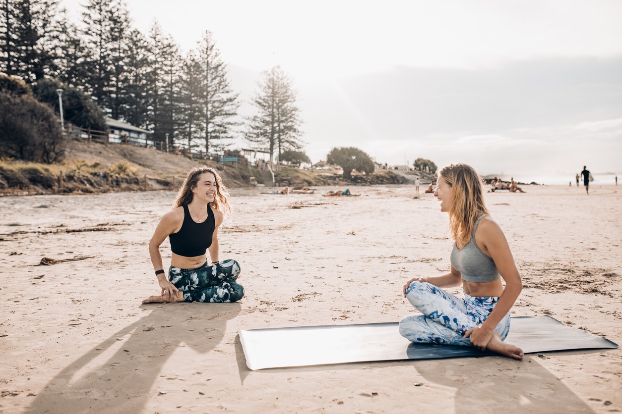 Two women perform the cow face yoga pose on the beach. Noosa Vs Byron Bay