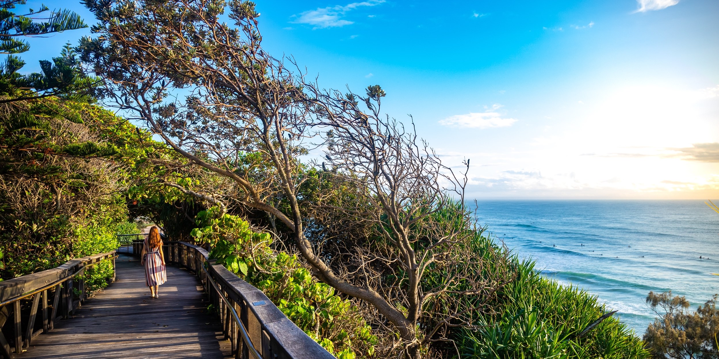 A girl in dress walks along the boardwalk and enjoys the view of the stunning Coolum Beach and surfers catching big waves, Pacific Ocean. Stunning panorama of Sunshine Coast, Queensland, Australia